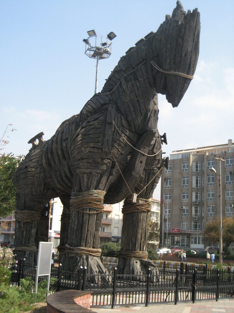 A large wooden replica of the Trojan Horse, located in the city of Çanakkale, Turkey. The structure is dark brown with rough, weathered planks of wood, bound together with thick ropes around its legs and body. The horse is positioned outdoors in a fenced area, with buildings and streetlights visible in the background.