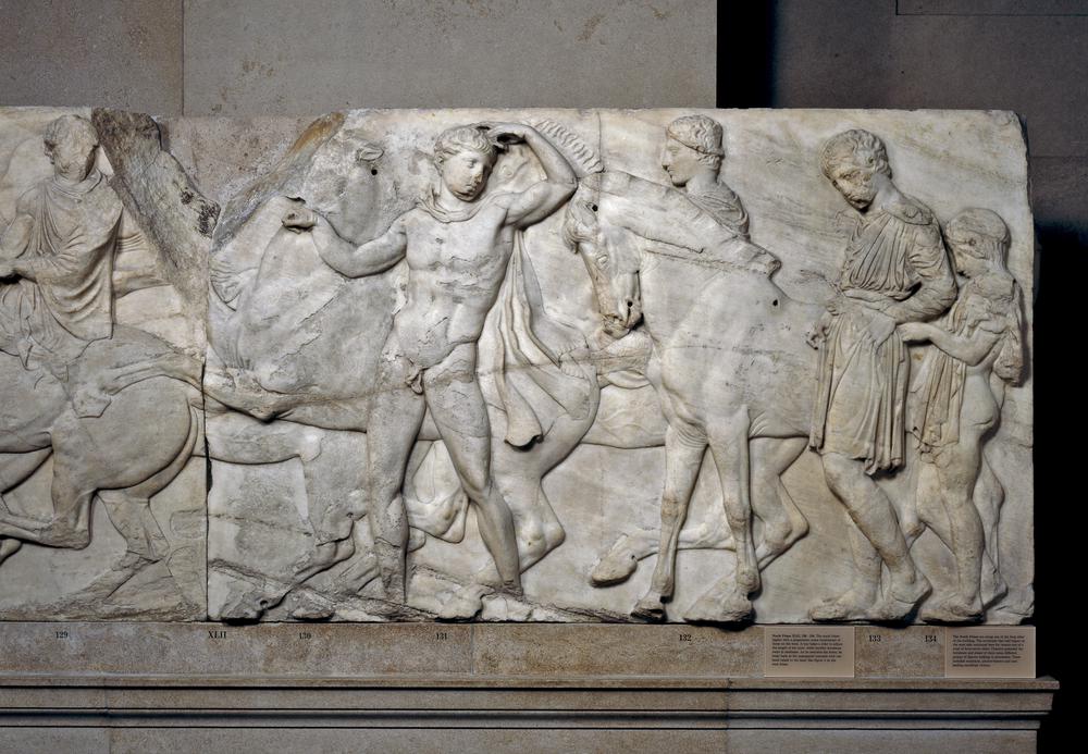 A marble relief from the Parthenon, depicting a dynamic scene of ancient Greek figures in intricate detail. The relief showcases several human figures engaged in a procession, characterised by flowing drapery and expressive poses.