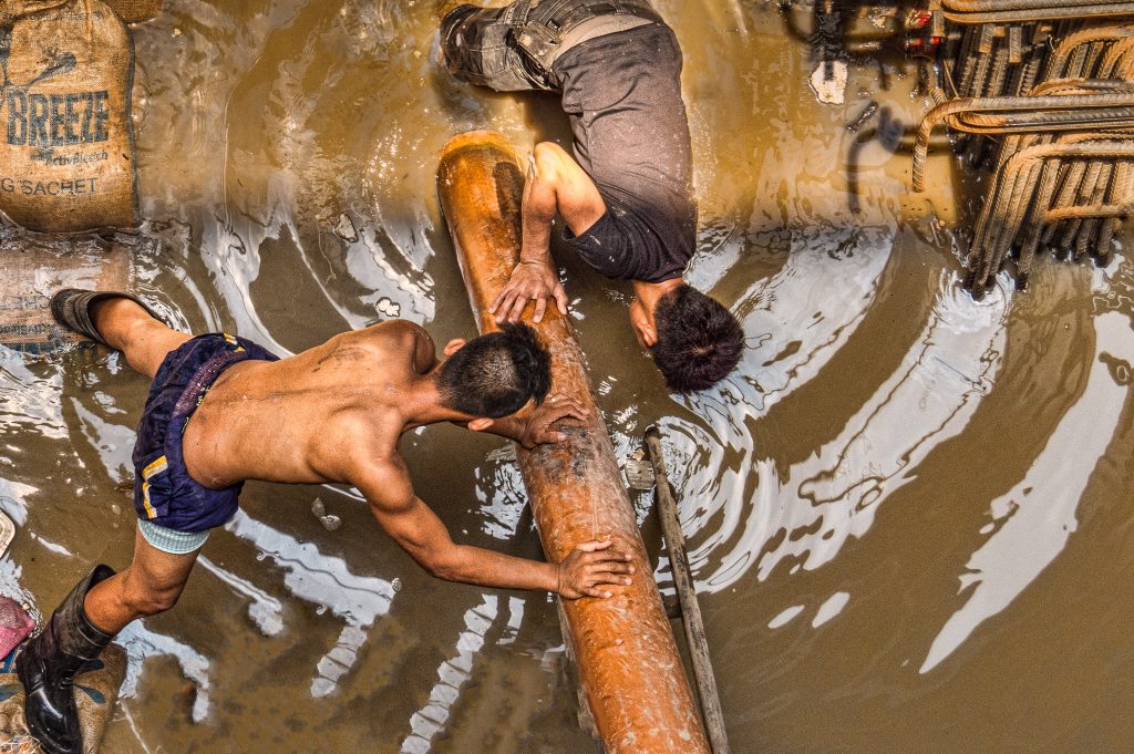 Two men are waist-deep in muddy water manually moving around a large timber log. They are surrounded by other building materials including steel rio which is used in supporting concrete. They are not wearing any type of protective clothing (one even doesn't have his shirt on) and they have no machinery assisting them.