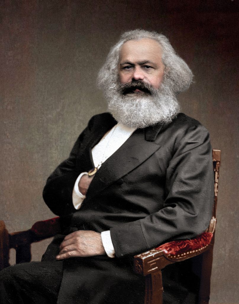 A portrait painting of Karl Marx from 1875. He is middle aged with a sizable white beard, a receding hairline and white hair. He is seated in a timber chair and is wearing a black suit jacket with a white shirt.