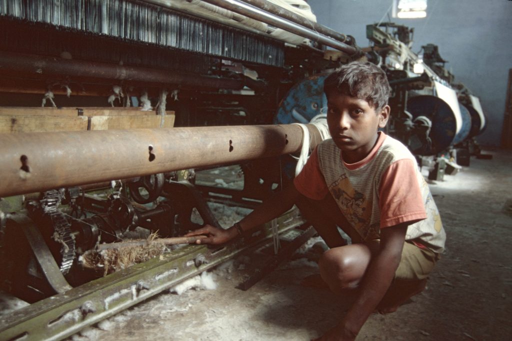 A young boy, approximately 12 years old Parkistani boy is cleaning what looks like textile mill equipment. The photo was taken in 2016 and clearly is a modern image of child labour.
