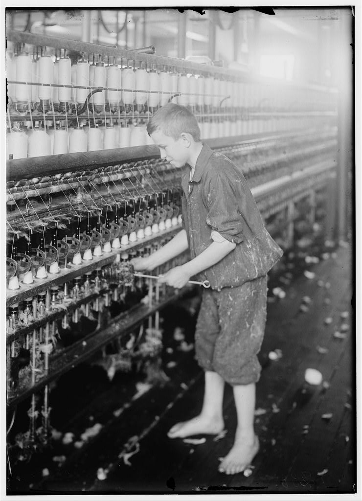 A 1916 photo in black and white depicts a young man cleaning a cotton spinning machine. He is bare foot and covered with cotton dust.