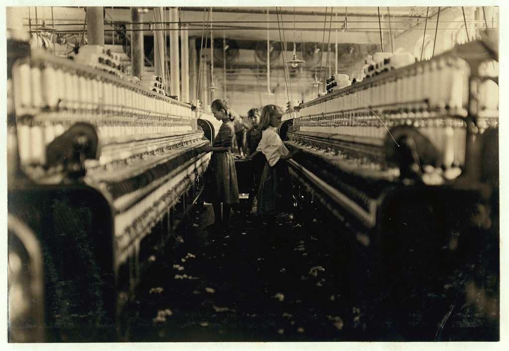 The images looks down an aisle between industrial cotton machines. We see two girls of around 10 - 12 years old working and another child, perhaps a boy, also carrying out a work task.