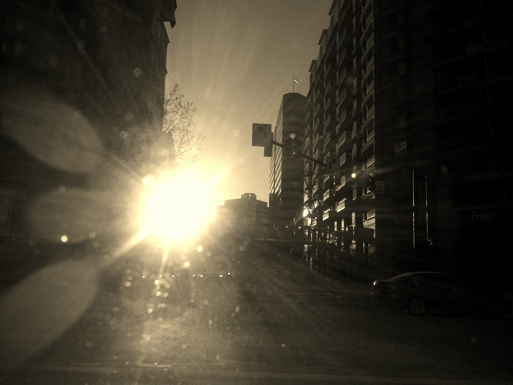 The rising sun causes glare on a dirty windshield in an urban street. The image almost appears to be in black and white.
