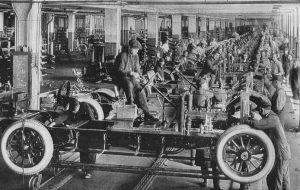 A line of cars are at varying levels of assembly. They are all sitting on rails that move each car from one team of workers to the next. This image is in black and white and is from 1923 in America.