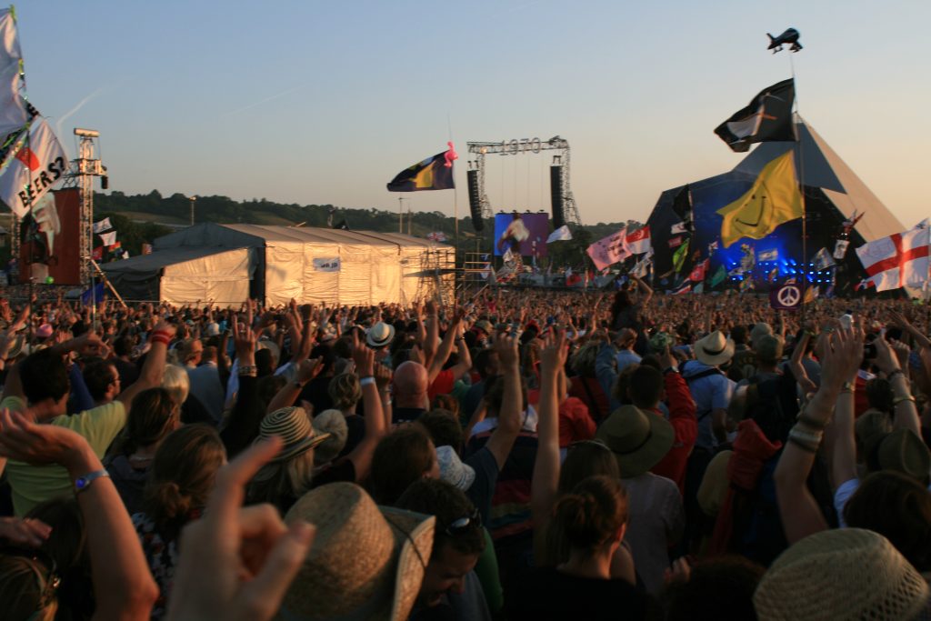 A crowd of thousands look towards a performance on a large stage. There are many different flags flying ranging from different countries to a smiley face icon. Many of the crowd have there hands up.