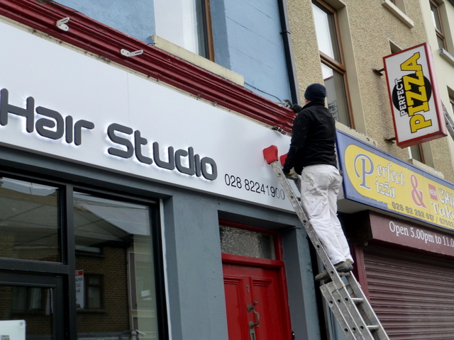 A commercial painter is up a ladder painting the exterior of a hair dresser shop.