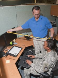 A man is conducting an ergonomic analysis for a woman sitting at a computer workstation. He is measuring the distance between her computer monitor and her head.