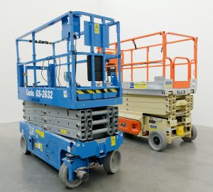 This image is of two large scissor lifts with baskets with safety rails. They are not in use as their scissor like lifting arms are clasped down for storage.