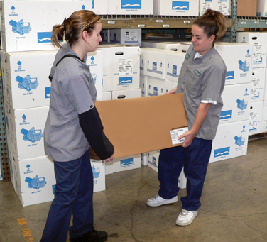 Two women workers are collaborating to lift a box in a warehouse setting.