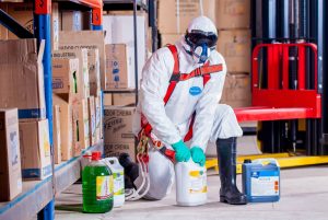 A worker in full protective equipment including a whole jumpsuit, gloves, face mask and googles is handling chemical containers on the floor in an industrial warehouse setting.