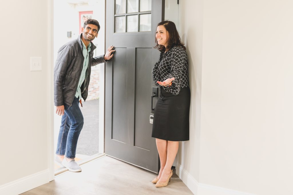 A female real estate agent is standing at an open front door welcoming in a male client. Both are very happy and comfortable in this situation.