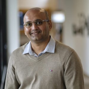 Image of a male wearing glasses, a blue shirt and a light brown jumper.