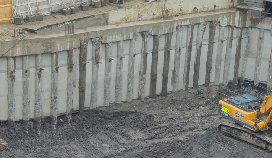 Construction site featuring a deep excavation with reinforced concrete piles arranged in a row and connected with a pile cap. Anchors are installed below the pile cap.