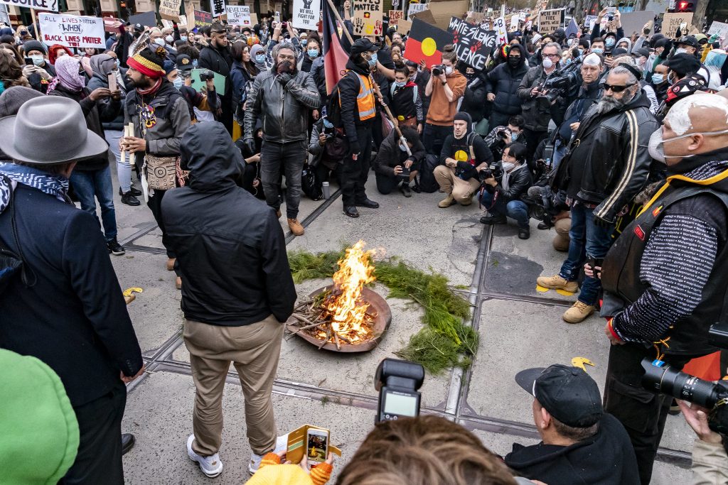 Group of people standing and sitting around a fire at a Black Lives Matter protest. Many of the people are wearing masks and holding phones, cameras, banners and protest signs.