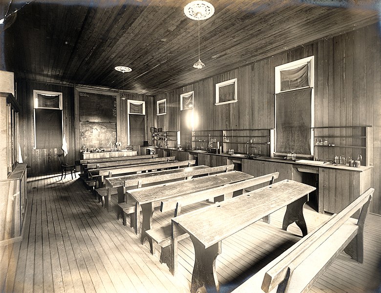A very simple wooden classroom. There are bench seats and the windows are blocked.