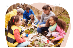 Three teachers and five children are in the middle of a lush green forest. They surround a long white rectangle board that has lots of items they have discovered in the forest. There are leaves, deer antler, tree bark and moss.