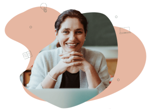 Female teacher looking at the camera sitting at her desk in the classroom. She has her hands linked and a laptop in front of her