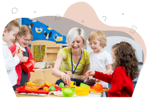 A smiling teacher sits at a table with four primary school aged children in school uniform. The teacher is guiding the children in play. They are all interacting with the fake food and drinks.