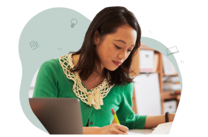 A teacher sits at her desk wearing a green cardigan. She is writing something down on a piece of paper.