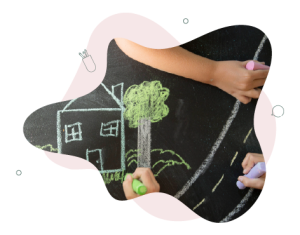 Child's hand holding a large piece of pink calk. They are drawing on a black board and adding to an image that has already been drawn (in chalk) of a road and houses.