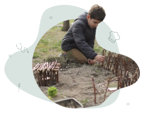 A young boy in a blue jacket is kneeling on the ground outside. He is using sticks he has found to build a house and a fence