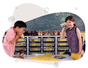 Two children standing in a classroom facing each other. They have two tin cans connected with a string. One child is speaking into the can whilst the other is listening and laughing