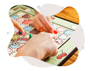 Two children's hands and one adult hand are pointing to a maths game. One of the children is holding a red game piece and is about to place it on the game board with the help of the teacher.