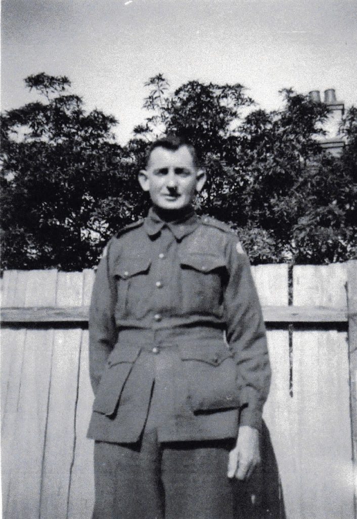 Black and white photo of Jim Wiggins in his Army uniform. He stands in front of a wood picket fence with trees in the background.