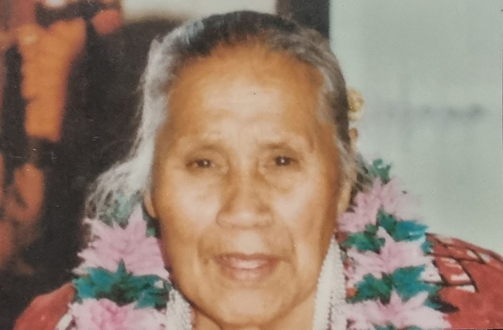 A close-up photo of an older person with gray hair pulled back, wearing a colorful pink and green flower lei and a red patterned garment, with a blurred background.