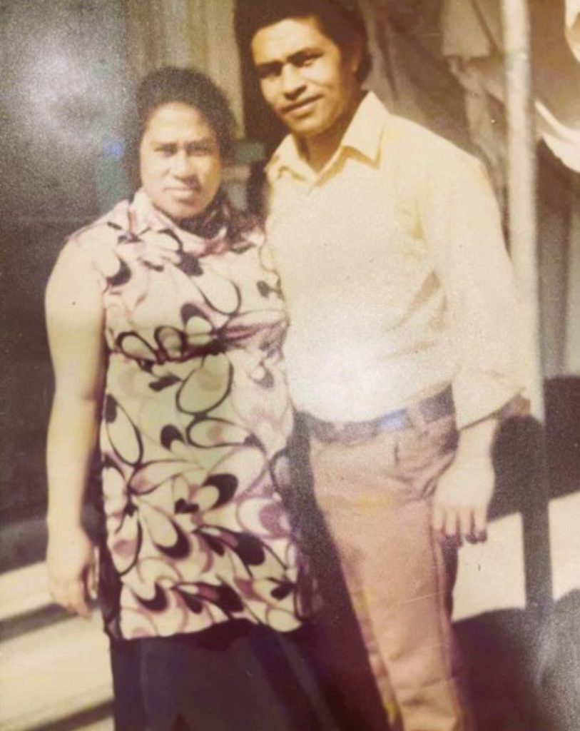 A vintage photo of two people standing together: one in a sleeveless patterned top and dark pants, the other in a light shirt and light brown pants, both facing the camera.