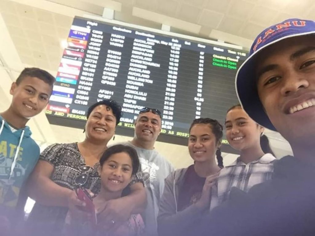 Six people stand together smiling at the camera in an airport, with a large flight information board behind them displaying flights to cities like Los Angeles, Auckland, and Honolulu.