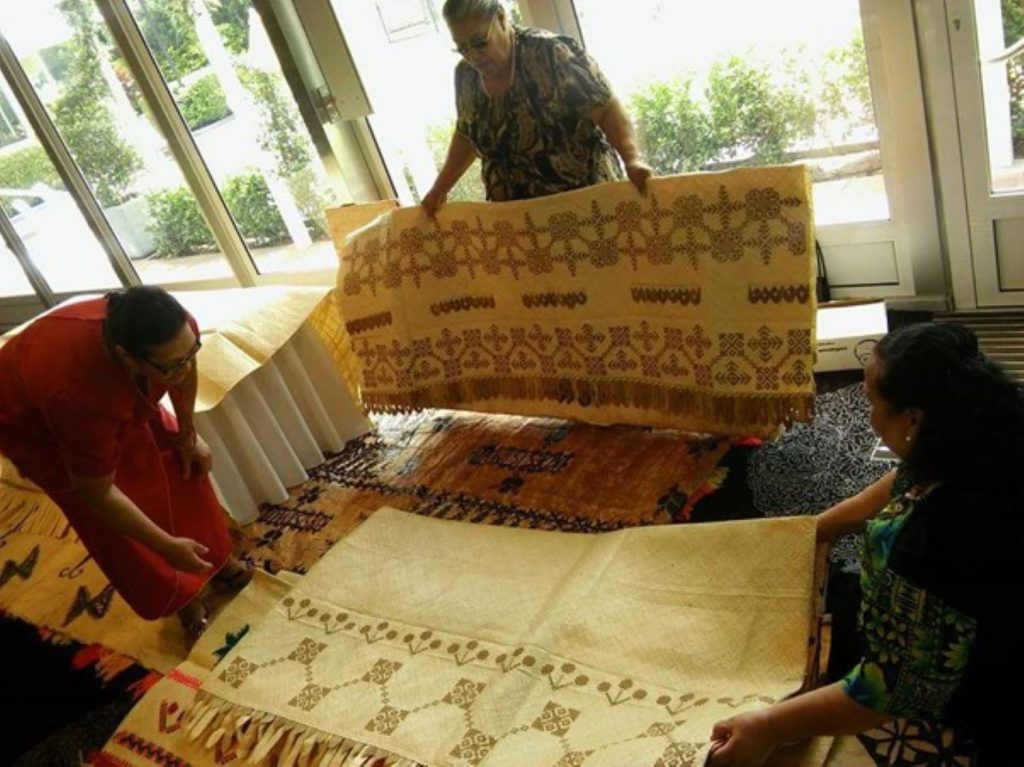 Three people examine traditional woven mats in a room with large windows. One holds up a mat with geometric and floral patterns, while others closely observe the detailed designs and fringes.