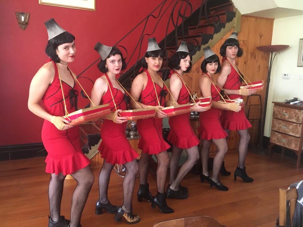 Six people in matching red dresses, black fishnet stockings, and gray hats stand in a line indoors, each holding a tray, with a staircase and red walls behind them.