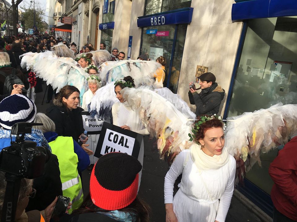 A group of people in white costumes with feathered wings and green wreaths protest on a city street, holding signs reading “COAL KILLS” and “Free Our Power.”