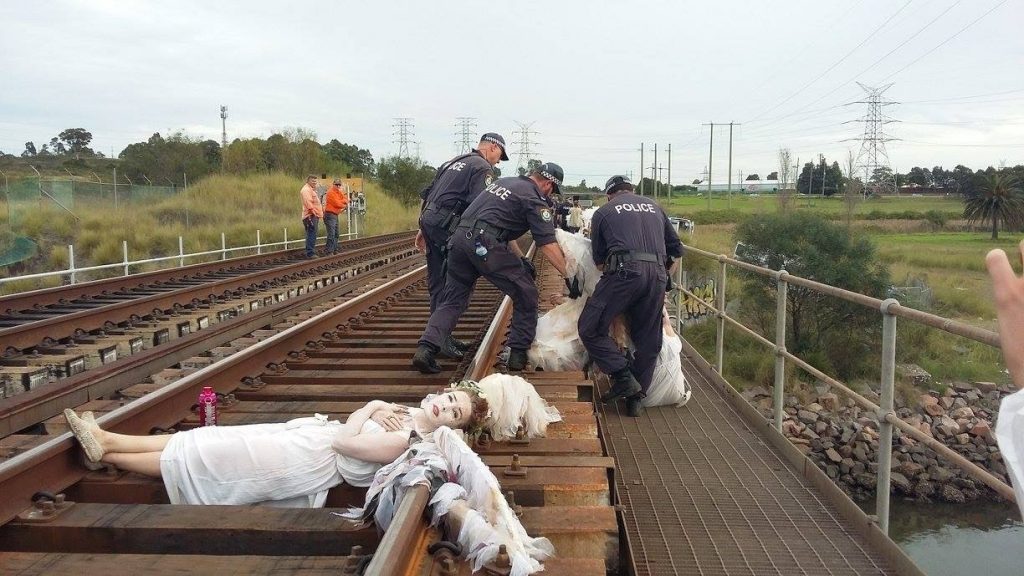 A group of police officers on a railway bridge detain people in white angel-like clothing lying on the tracks. There is grassy fields and power lines in the background.