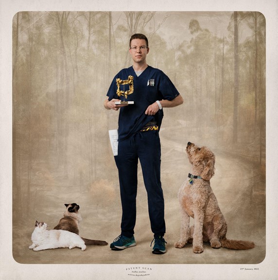 Tall young man standing against a backdrop of the Australian bushland with his two cats and dog sitting at his feet looking up at him.