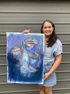A picture of a amiling Filipino woman with shoulder length dark hair, wearing blue shorts and a gray t-shirt and holding onto the final painting that formed the cover of this textbook. Please see the "About the Cover" section of this book for a detailed description of the cover art.