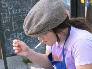 A White woman with brown hair in a newsboy cap and wearing a lilac shirt and blue apron is sitting outside a coffee shop, in front of a chalk board, smoking a cigartte.