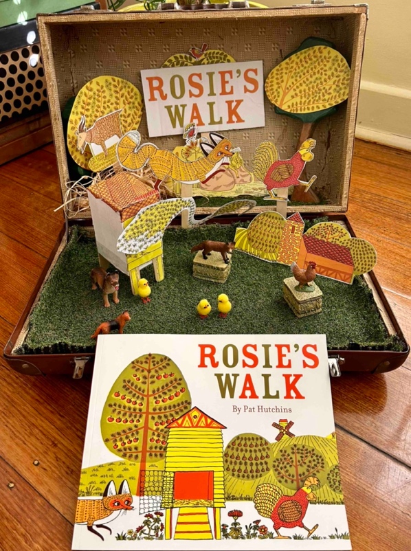 Cardboard cut-outs of illustrations from the picture book, Rosie's Walk, are presented on a small patch of fake grass, with some small toy farm animals.