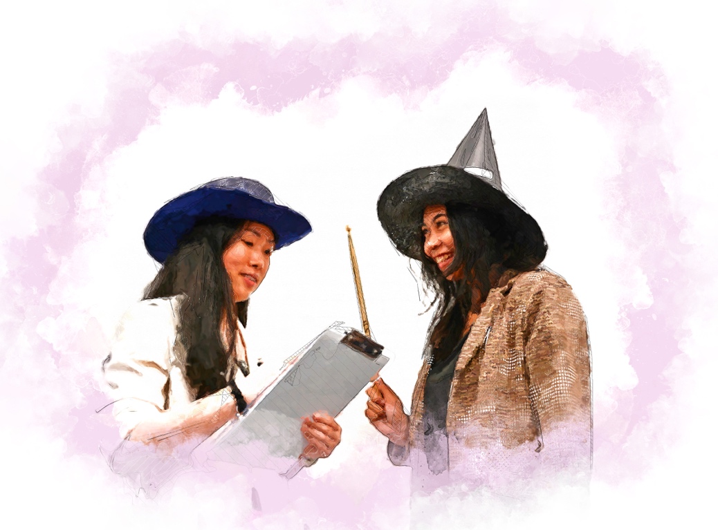 Yuwen is wearing a blue cowboy hat and writes something on a clipboard. Charlotte stands next to her wearing a witches hat and holding a wand.