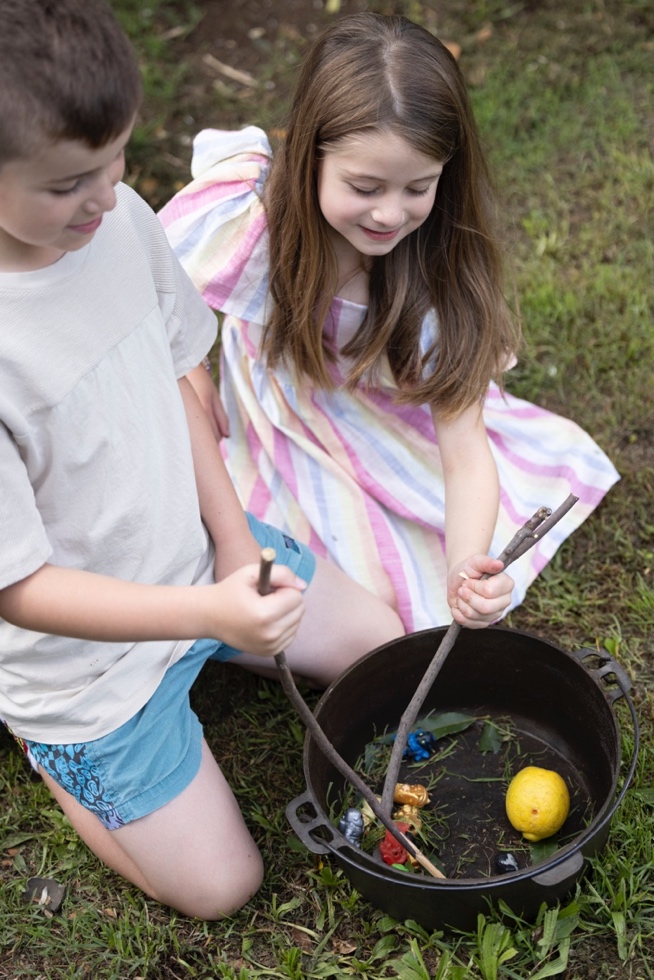 A young boy and girl kneel next to a cooking pot which sits on some grass. The stir the 'ingredients' a lemon, some grass and other items.