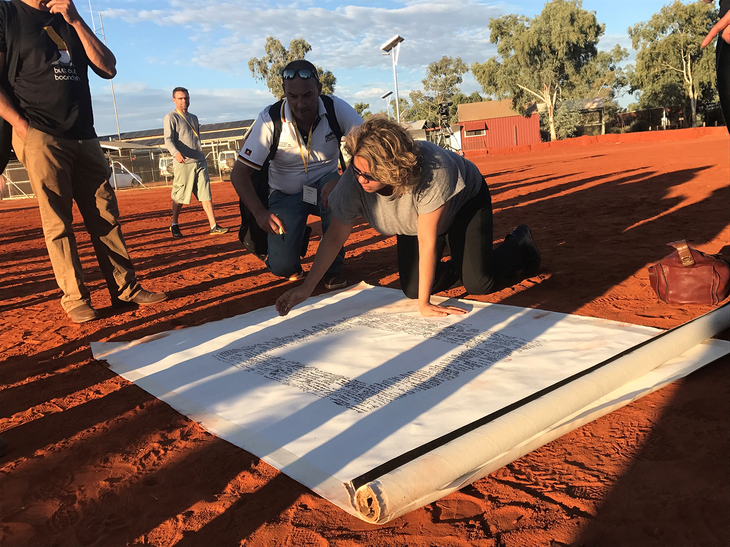 CEO of the Yothu Yindi Foundation, Denise Bowden, kneels on the ground to sign the Uluru Statement from the Heart, watched by several other people. The Uluru Statement is a large painted canvas, spread out on the red dirt.