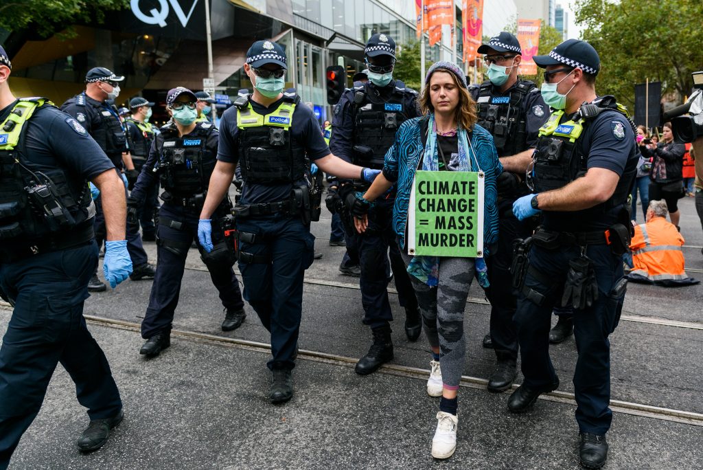 Photo of climate protestor being arrested in Melbourne, Australia. The protestor has a sign hanging around their neck that reads "CLIMATE CHANGE = MASS MURDER". There are six police officers surrounding the prosestor.